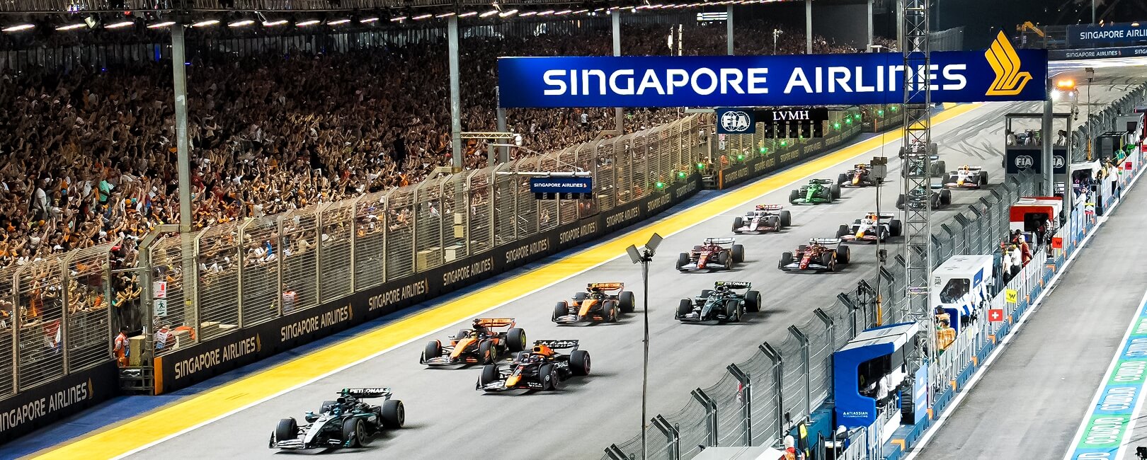 Formula 1 cars racing under lights past a packed grandstand at the Marina Bay Street Circuit.